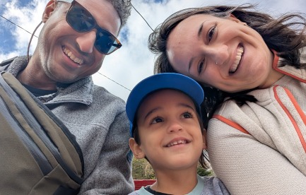 Family of 3 smiling with a blue sky and clouds in the background