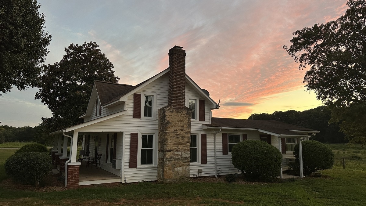 White farmhouse with red trim in front a colorful evening sky