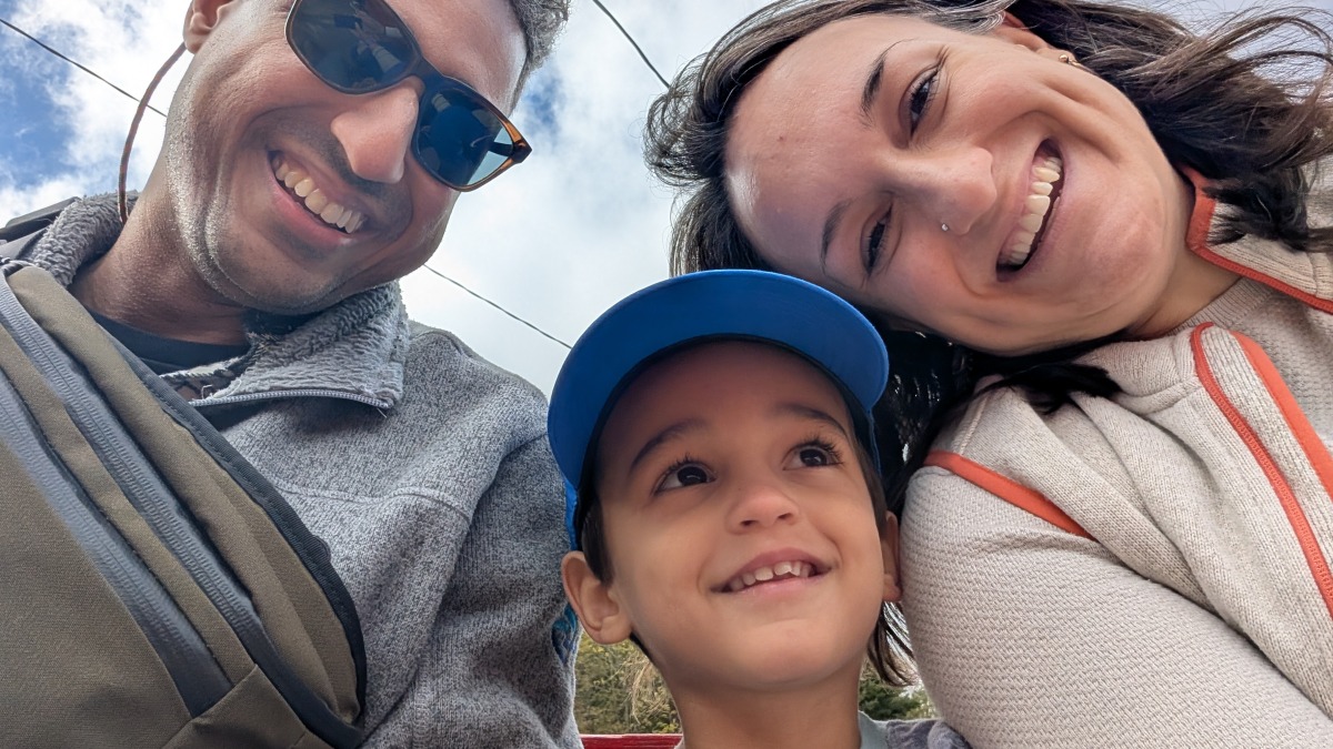 Family of 3 smiling with a blue sky and clouds in the background