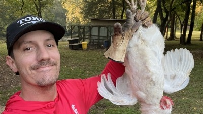 A man in a red shirt and black cap stands outdoors holding a white chicken by its legs. Trees and a small coop are visible in the background, showing a natural farm setting and humane livestock management.
