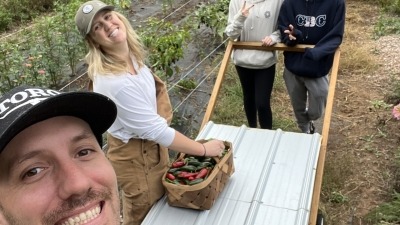 A smiling group of four stands together in a lush vegetable garden. One person holds a cart with a basket of freshly harvested peppers. The scene reflects teamwork, joy, and hands-on connection to growing food.