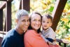 Family of three smiling for a photo with a bridge and autumn leaves in the background.